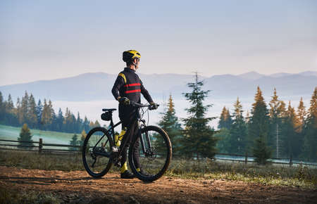 Male cyclist standing with bike with coniferous trees and hills on background. Man bicyclist enjoying bicycle ride in mountains in the morning. Concept of sport, biking and active leisure.の写真素材