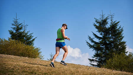 Trail runner running up to meadow outdoors under blue sky and looking at his watch. Jogging in open nature space in sunshine warm day. Concept of sport, training and active leisure.の写真素材