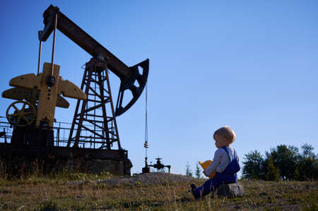 Rear view of preschool guy sitting on one stone, holding construction helmet in hands. Male kid looking at operation of drilling rig station for oil extraction outdoors under blue sky.の写真素材