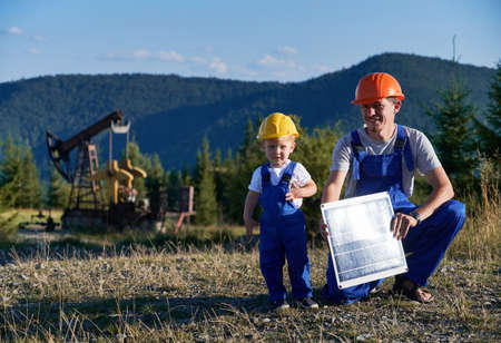 Portrait of professional Caucasian mechanic with solar panel learning capable boy derrick maintenance, father and son dressed in blue uniform looking at camera during daytime at petroleum stationの写真素材
