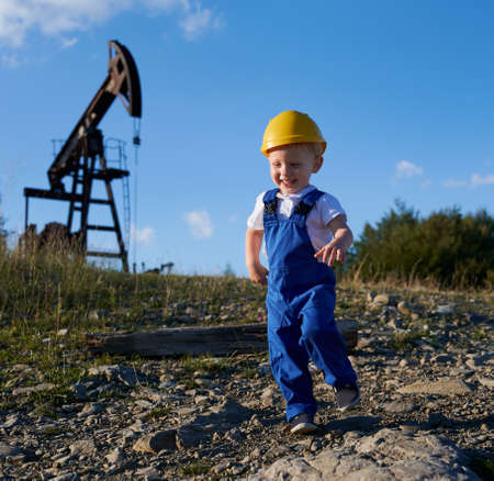 Happy preschool boy in work uniform and construction helmet running on stone road outdoors on the background blue summer sky and oil well at left. Equipment for oil industry.の写真素材