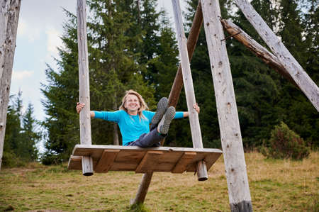 Happy woman sitting with straight legs on wooden seat attached to large wooden poles which she...