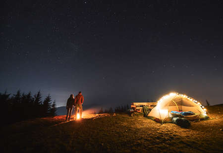 Young man and woman travelers holding hands while standing near bonfire under night sky with stars. Beautiful view of night starry sky over grassy hill. Concept of hiking, night camping, relationshipsの写真素材