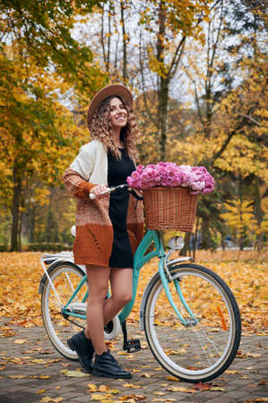Pleasant bike ride along alleys of autumn park. Female beautiful bicyclist standing near retro street cycle with basket, filled by pink flowers at yellow town forest.の写真素材