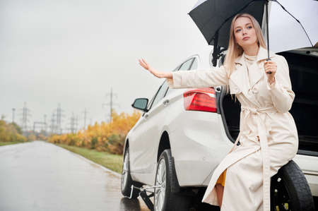 Beautiful woman on road near her white car with punctured car tire. Female driver asking for help, holding umbrella, sitting on spare wheel.の写真素材