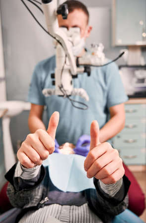 Close up of young woman showing approval gesture while lying in dental chair. Male dentist checking female patient teeth while woman giving thumbs up. Concept of dentistry, dental care and approval.の写真素材