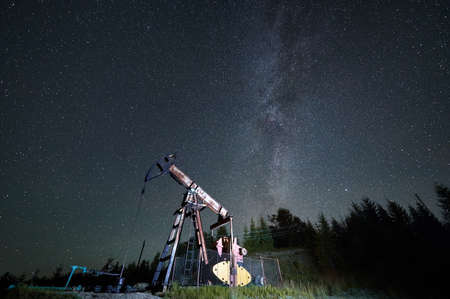 Against the background of magical starfull sky and Milky Way galaxy, oil rig in process of petroleum extraction. Oil production technology by pumpjack outdoors in oilfield.の写真素材