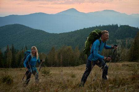 Young woman with glad smile on face following by her husband on hiking route while traveling on mountain spaces. Boundless woods and mountain ranges under the evening sky on the background.の写真素材
