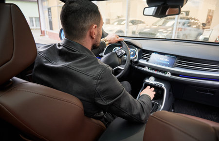 Back view of handsome young man choosing new car at dealership, sitting inside a car.
