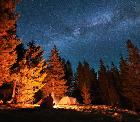 Rear view of couple in love relaxing in their camp at evening. Vacationers sitting by campfire and looking at the starry sky with Milky way. Romantic atmosphere during camping at starry night outdoorsの写真素材