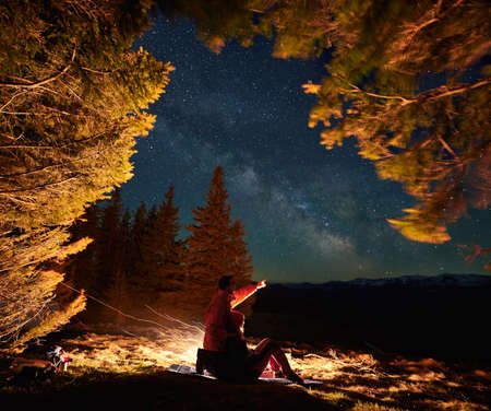 Side view of romantic couple in the mountains resting by campfire on tourist rug among meadow under...