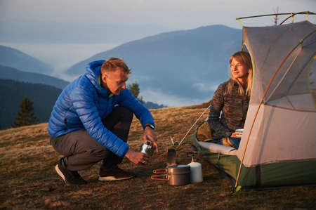 Male tourist making hot coffee outdoors at his campsite next to his wife, who sitting in tent and smiling tenderly at him against the backdrop of mountain hills. Concept of camping and relationships.の写真素材