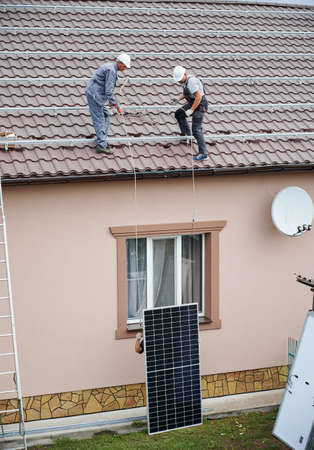 Men installers lifting up photovoltaic solar modul on roof of house. Electricians in helmets installing solar panel system outdoors. Concept of alternative and renewable energy.の写真素材