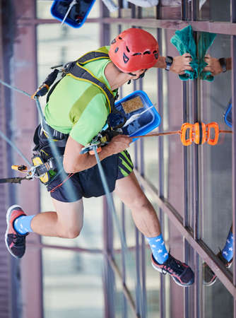 Industrial mountaineering worker washing glass windows of high-rise building, hanging on safety...