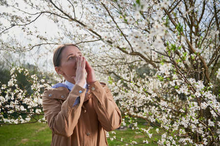 Woman allergic suffering from seasonal allergy at spring, posing in blossoming garden at springtime. Young woman sneezing in front of blooming tree. Spring allergy conceptの写真素材