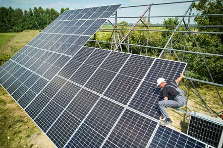 Male worker in safety helmet installing photovoltaic solar panel on sunny day in grassy field. Man...