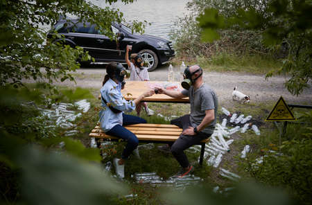 Man and woman in gas masks sitting on bench and toasting with plastic cups while spending time with...