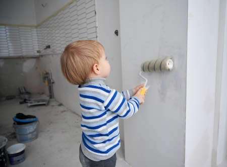 Cute little boy painting wall in room under renovation. Adorable child in striped sweater using paint roller while renovating apartment. Concept of home renovation, restoration and childhood.の写真素材