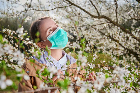 Woman allergic suffering from seasonal allergy at spring, posing in blossoming garden at springtime, wearing medical mask among blooming trees. Spring allergy conceptの写真素材