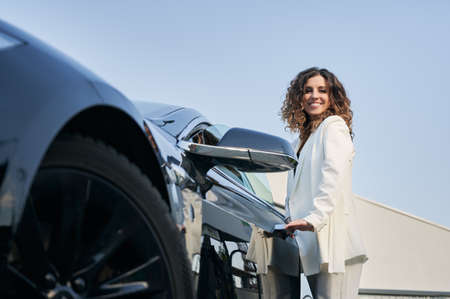Bottom view of happy smiling business girl with curly hair standing outside car and easily opening door of her auto. Beautiful woman holding handle on car door on the background of blue sky.の写真素材