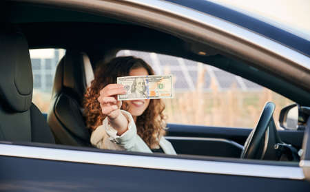 Close up of young woman with 100 dollar banknote in her hand sitting in electric vehicle....