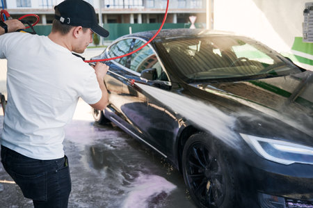 Male driver in white shirt using high pressure water sprayer while washing modern electric car on the street. Young man cleaning his vehicle at self-service car washing station.の写真素材