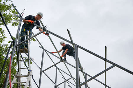 Man in safety helmet standing on ladder and holding hammer while colleague installing metal rail for solar modules. Male workers assembling support structures for solar panels under white sky.の写真素材