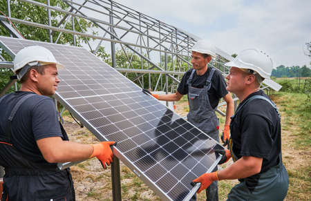 Three men solar installers building photovoltaic solar panel system outdoors. Male workers wearing...