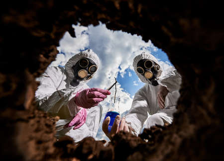 Two laboratory workers in protective clothing, rubber gloves and respirators taking sample of soil content from the earth into flask with reagent for research. View from inside pit.の写真素材
