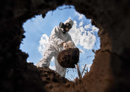 View from inside pit of ecologist digging pit by shovel and planting tree, wearing gas mask and...