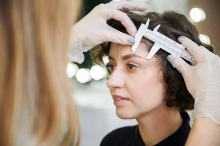 Esthetician using measuring tool for brow correction procedure in beauty salon. Close up. Beauty specialist placing ruler on woman forehead while taking measurements for eyebrow correction.の写真素材