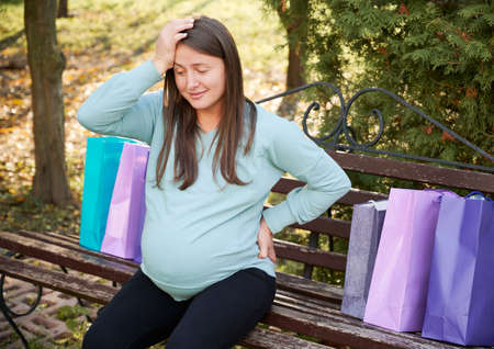 Surprised pregnant woman sitting on street bench, putting one hand on her forehead and other on waist. Expectant mother resting on wooden bench after shopping, putting packages next to each other.の写真素材