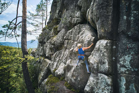 Senior alult woman climbing difficult route on a high boulder with rope. Fearless climber training...