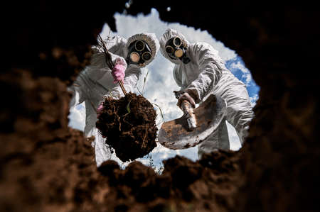 View from inside dug pit in which two people, completely protected from effects of hazardous substances, planting tree. Landscaping of dangerous contaminated territory.の写真素材