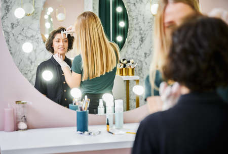 Young woman looking in the mirror and smiling while esthetician measuring eyebrow distance with brow ruler. Beauty specialist using measuring tool before eyebrow correction procedure in beauty salon.の写真素材