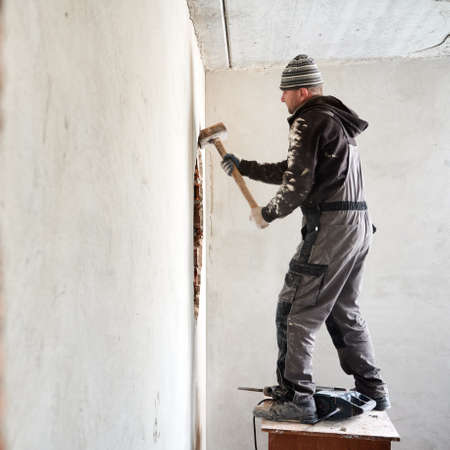 Experienced worker dismantling internal wall. Man standing on elevated surface, where perforator located, and with help of sledgehammer increasing through hole in the wall.の写真素材