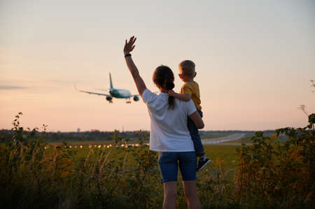 Back view of mother with toddler waving hands to landing commercial airplane at sunset. Lifestyle and travel concept.の写真素材