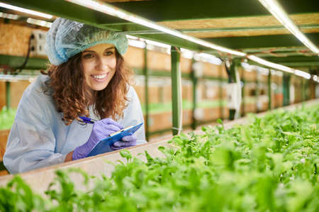 Female researcher in disposable cap looking at green leafy plants and smiling while writing on...