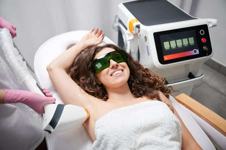 Young woman in safety glasses lying on daybed and smiling while beautician removing hair from female armpit with laser device. Cosmetologist performing laser hair removal procedure in beauty salon.の写真素材