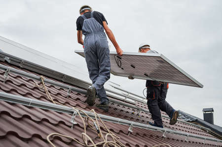 Men technicians carrying photovoltaic solar moduls on roof of house. Builders in helmets installing solar panel system outdoors. Concept of alternative and renewable energy.の写真素材