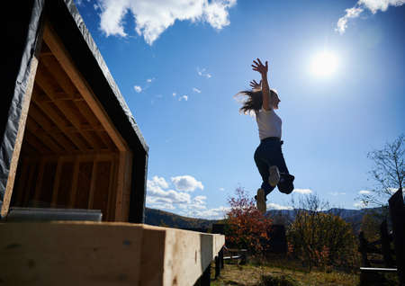 Happy woman on construction site jumping from terrace at unfinished wooden frame house on sunny day.の写真素材
