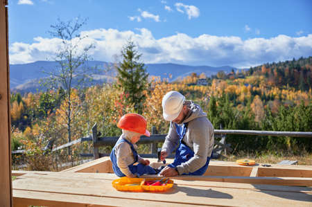 Father with toddler son building wooden frame house. Male builders hammering nail into plank on construction site, wearing helmet and blue overalls on sunny day. Carpentry and family concept.の写真素材