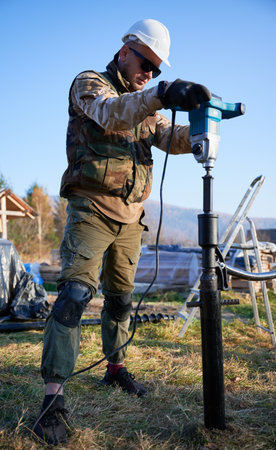 Male worker building pile foundation for wooden frame house. Man builder in white safety helmet drilling piles into the ground on blue sky background.の写真素材