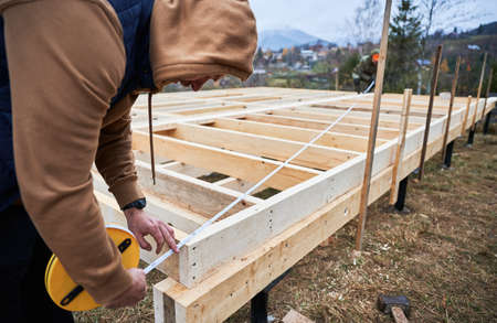 Man worker building wooden frame house on pile foundation. Carpenter using tape measure for...
