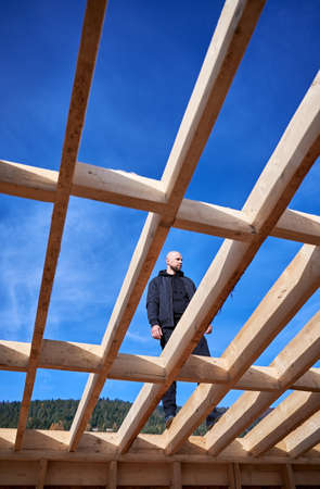 Male carpenter building wooden frame house. Bald man standing on construction site, inspecting...