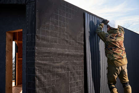 Male builder installing black corrugated iron sheet used as facade of future cottage. Man worker building wooden frame house. Carpentry and construction concept.の写真素材