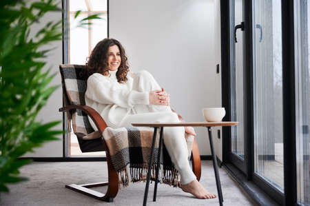Curly young woman resting inside contemporary barn house. Happy female tourist sitting on chair,...