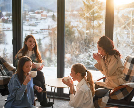 Young women enjoying winter weekends inside contemporary barn house. Four girls having fun and drinking hot tea near panoramic windows at sunset.の写真素材