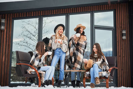 Young women enjoying winter weekends on terrace of contemporary barnhouse in the Scandinavian style. Four girls having fun, sitting on chairs and drinking hot tea.の写真素材