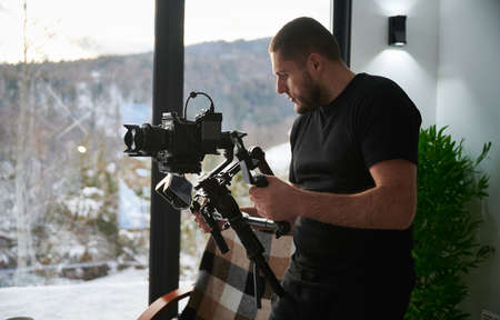 Videographer man shooting footage, using camera mounted on gimbal stabilizer equipment. Panoramic windows with winter landscape on background.の写真素材